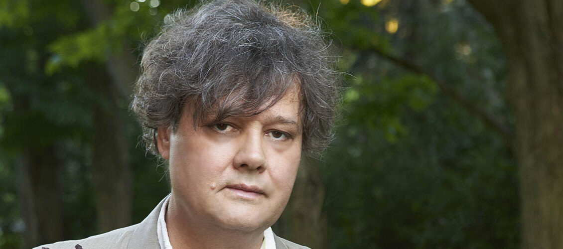 Ron Sexsmith is wearing a grey suit coat over top a striped shirt. He is standing in front of trees with green leaves.