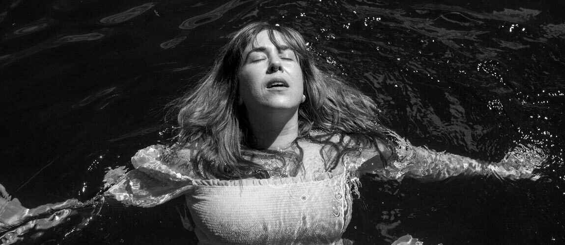 Serena Ryder wearing a white dress floating in water with her eyes closed by face looking at the sky. Photo is black and white.