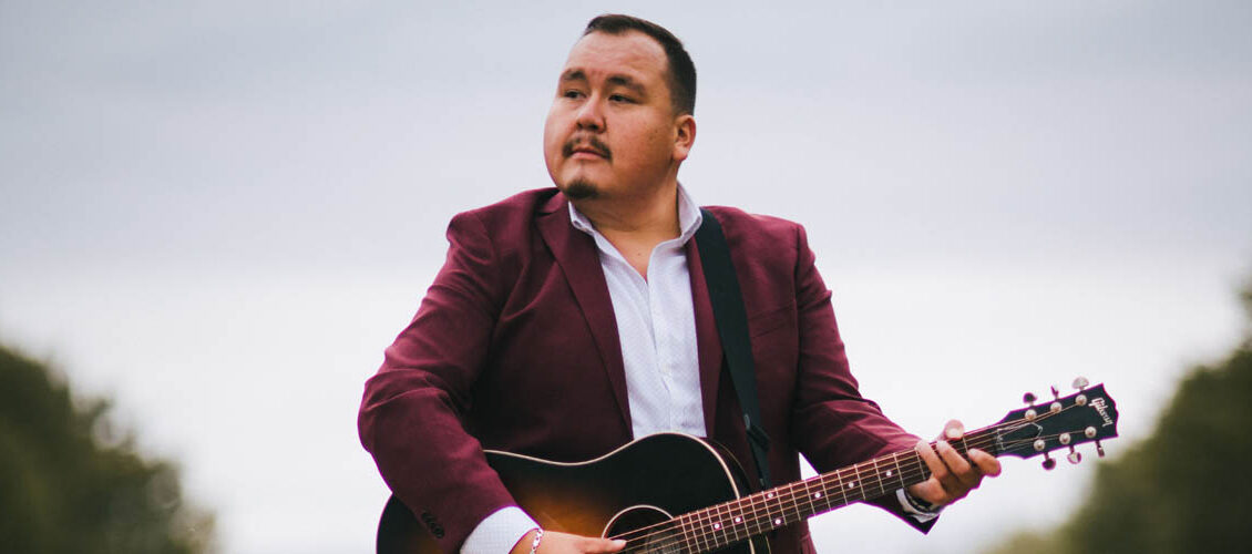 William Prince from the waist up, wearing a burgundy blazer and white shirt holding a guitar. Grey sky and tree tops in the background