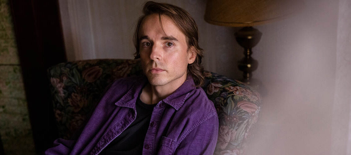 Andy Shauf looking at the camera with a few strands of hair in his face. He is wearing a purple button up shirt open over a black t shirt. He is sitting on a vintage floral print sofa in front of cream coloured striped wallpaper.