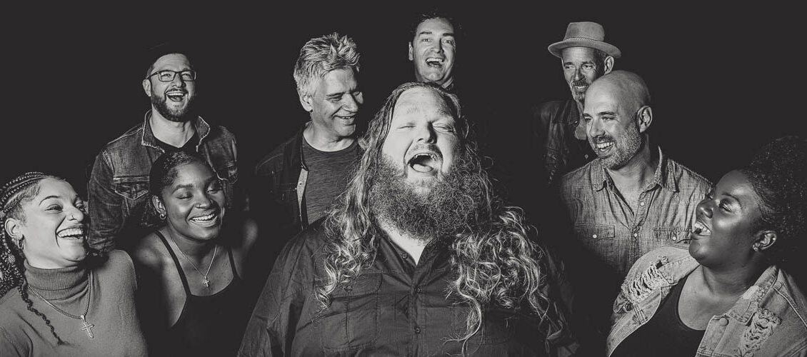 Black and white image of Matt Andersen singing surrounded by 8 people standing in two rows in a loose semi-circle with happy expressions on their faces as they look at Matt.