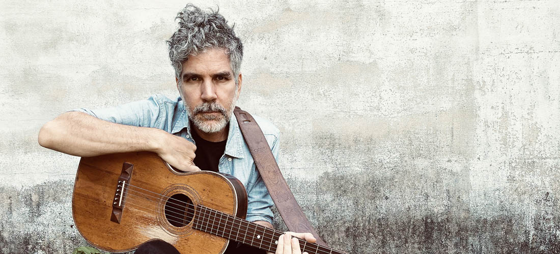 Hayden sits in front of a grey cement wall on a rough wooden bench leaning towards the camera with a serious expression. He has salt and pepper hair and beard, and is wearing a faded denim shirt and dark pants. He has his guitar on his knee and is leaning on it.