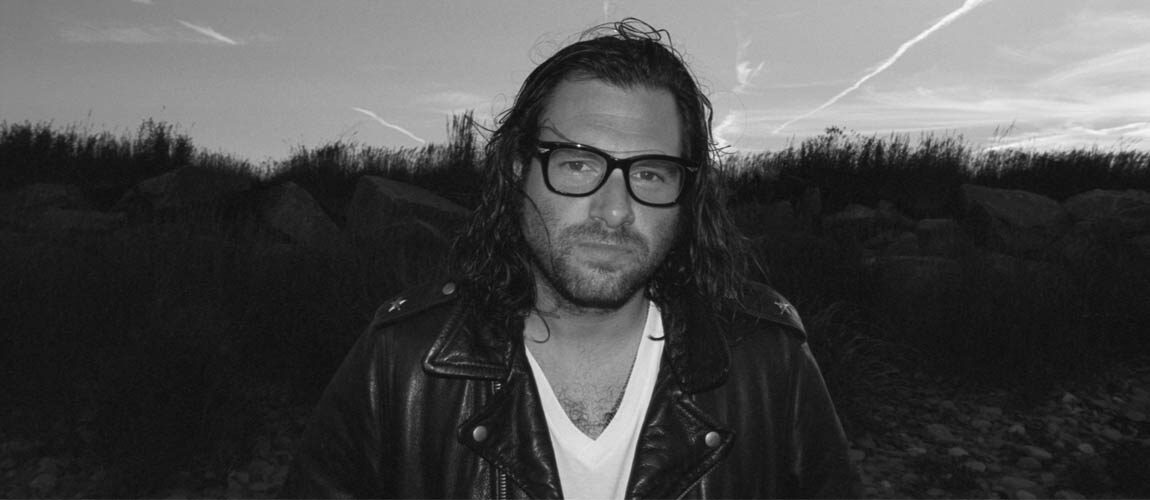 Black and white photo of Adam Baldwin. In the background, the bottom half of the image is dark trees, the top half is a grey sky. In the foreground, you can see Adam from the chest up. He is wearing a black leather jacket, white v-neck tshirt and some thick framed black glasses.