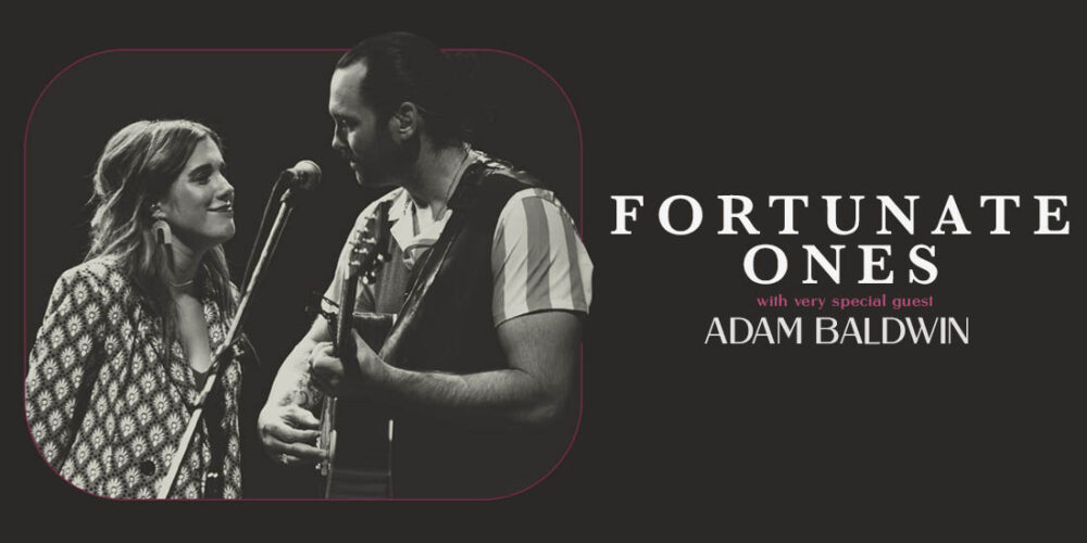 Black and white image of Fortunate Ones standing on stage looking at each other singing into the same microphone. Andrew is holding a guitar.