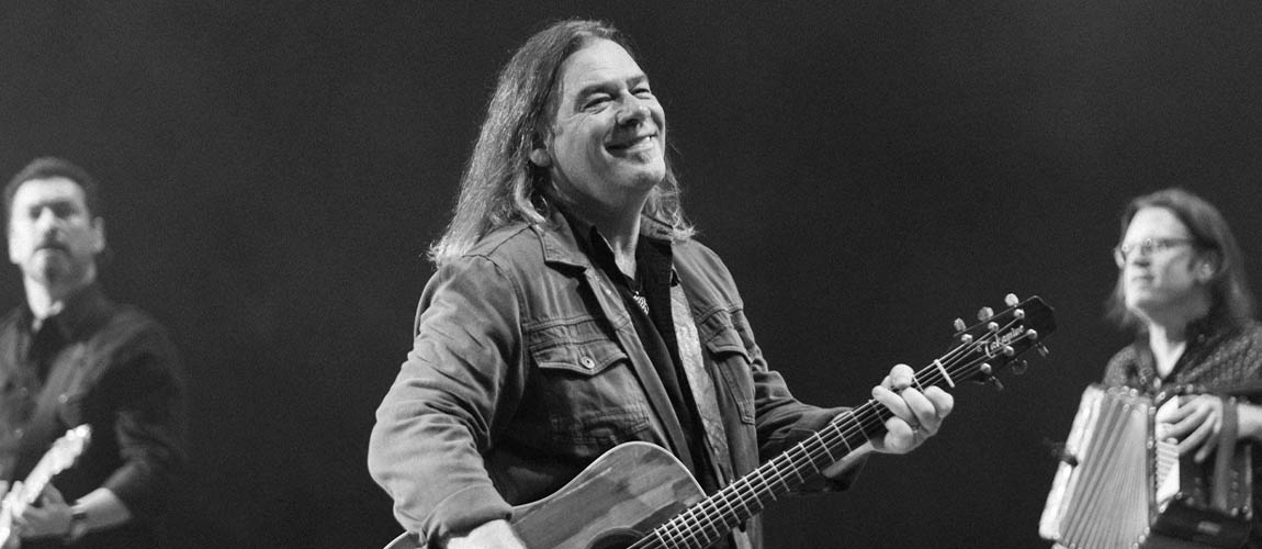 Black and white photo of Alan Doyle standing on stage wearing a jean jacket, dark pants, and playing guitar. He's looking off to the right smiling