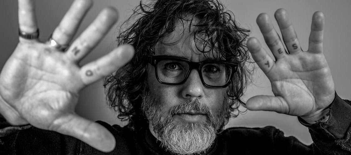 Kevin Drew is shown chest up in a black and white photo. He is wearing a black turtle neck. He has his hands up and open to the camera, with his palms facing the viewer. He has NO tattooed on two fingers and YES tattooed on the left hand. His hair is curly and he has a salt and pepper beard. He is wearing thick rimmed glasses.