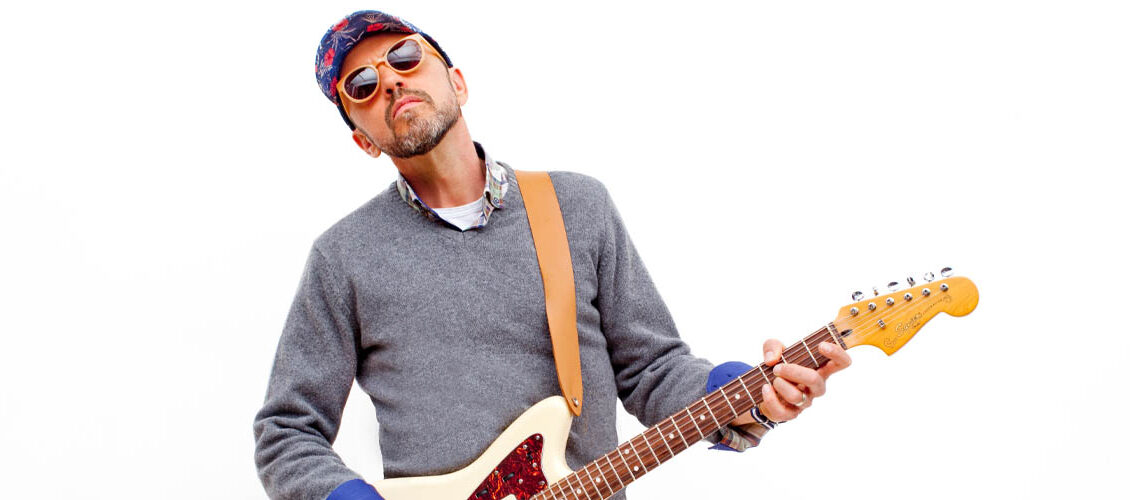 Hawksley Workman wearing a blue sweater, yellow pants, sunglasses, and ball cap. He's holding a white electric guitar.
