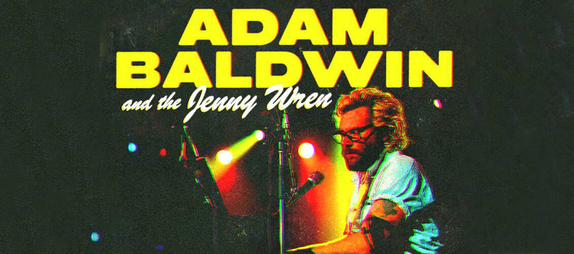 Adam Baldwin is sitting at a piano and singing into a microphone. He has bleached blonde hair, and is wearing glasses and a white short sleeved button up shirt. The text above his head reads Adam Baldwin and the Jenny Wren.