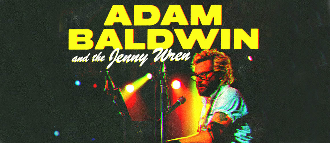 Adam Baldwin is sitting at a piano and singing into a microphone. He has bleached blonde hair, and is wearing glasses and a white short sleeved button up shirt. The text above his head reads Adam Baldwin and the Jenny Wren.