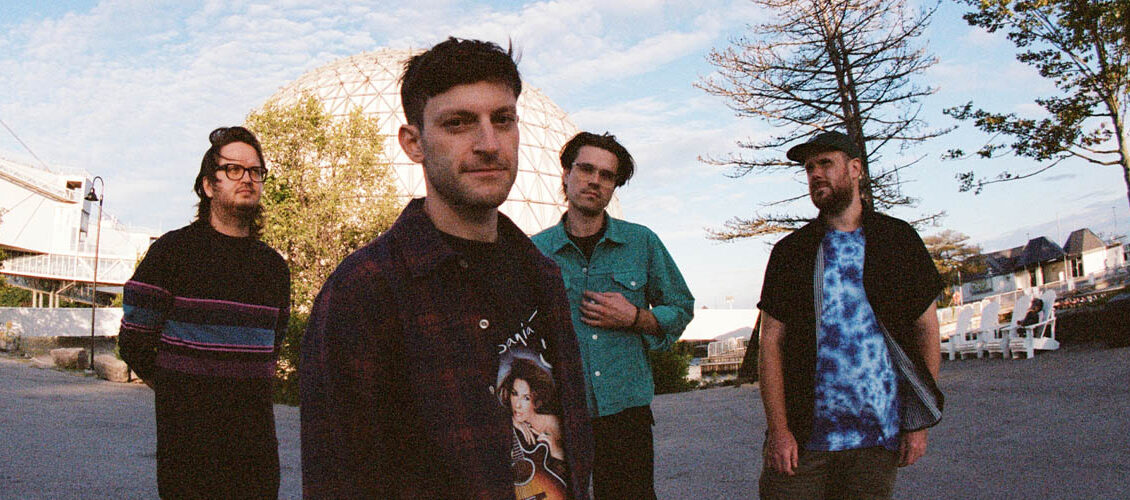 PUP is standing outside in a parking lot with sparse trees and a blue sky with white clouds in the background. The band is standing facing the camera. They're all wearing casual clothes.