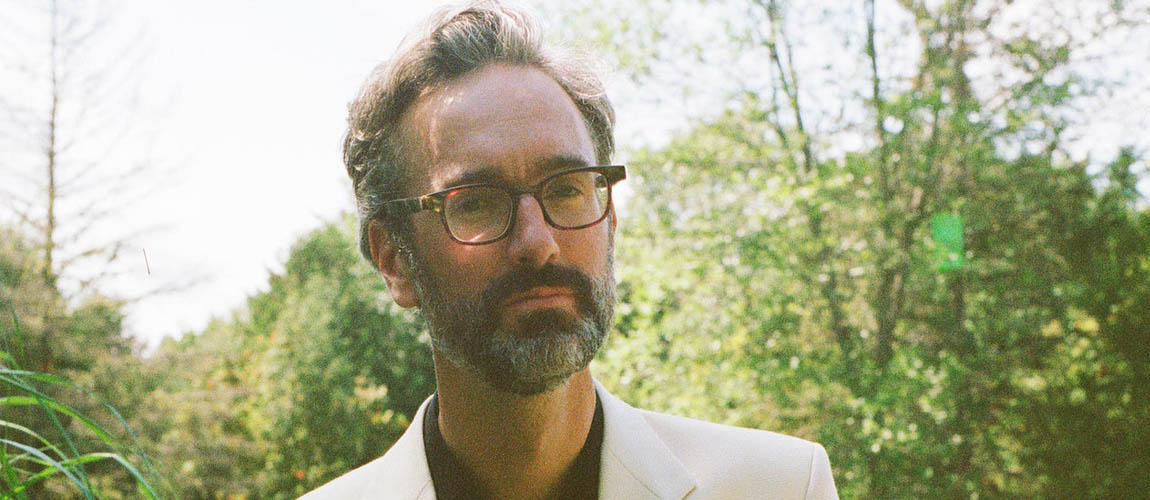 David Myles is shown from the shoulders up standing in front of leafy green trees on a cloudy day. He is wearing a white blazer and tortoise shell glasses. He has a serious expression on his face and is looking towards the camera.